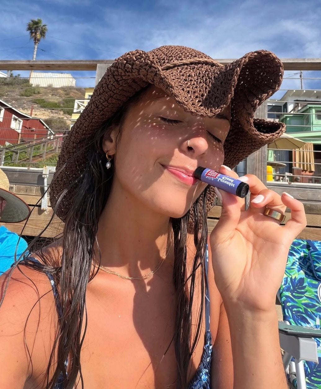 A woman outdoors wearing a brown crocheted cowboy hat and holding a Mood Indigo Lavender  lip balm tube near her lips. The background shows a sunny beach or coastal setting with boardwalk, a palm tree, and blue sky.