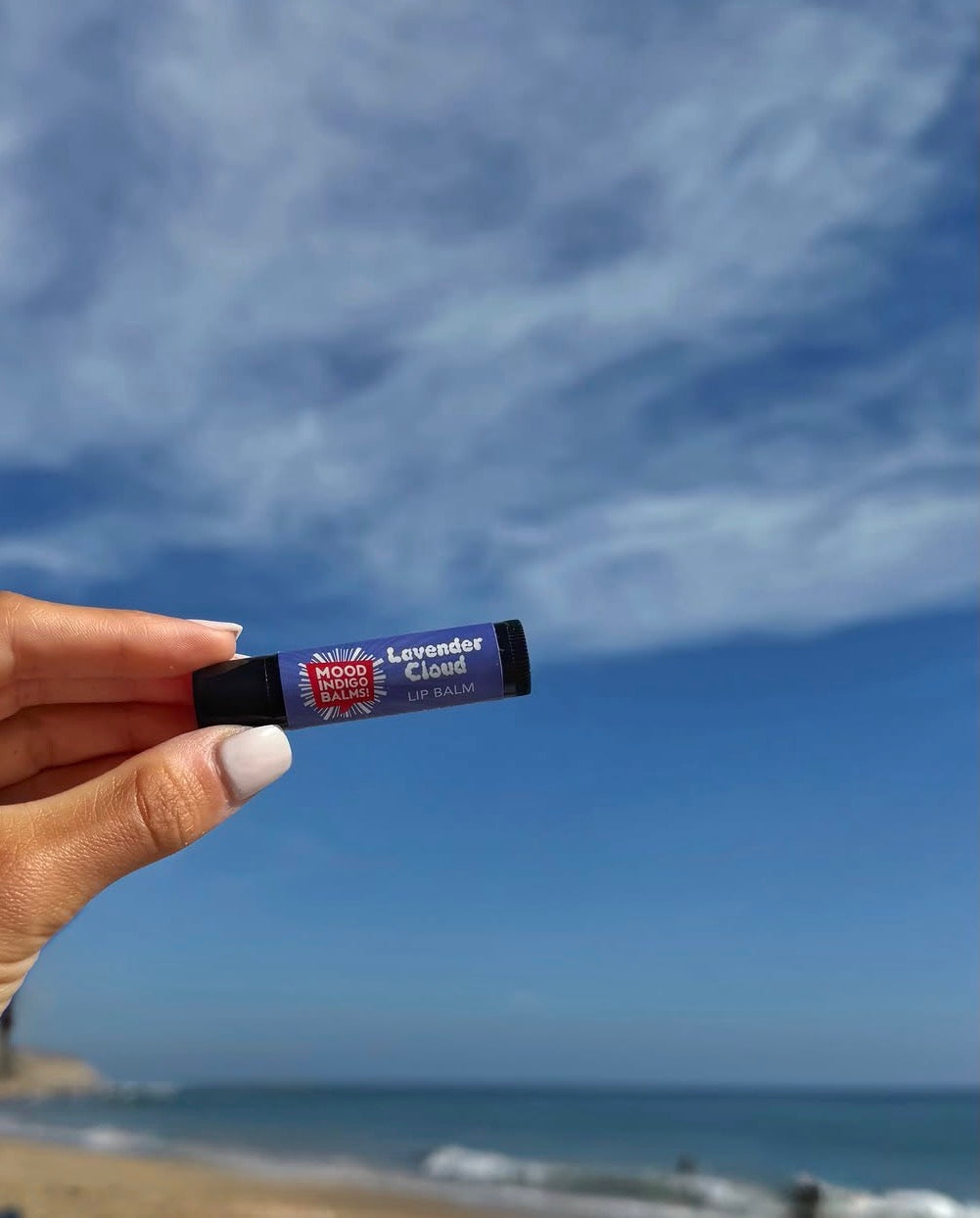 A hand holds a tube of Mood Indigo's all natural healing Lavender Cloud Lip Balm, against a blurred beach and blue sky background.