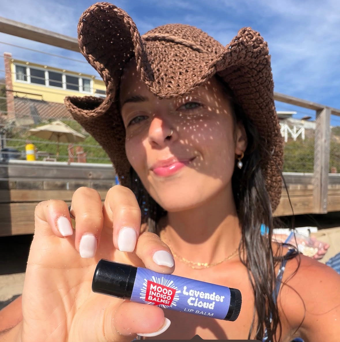 A woman outdoors wearing a brown crochet cowboy hat and holding a Mood Indigo lavender lip balm tube labeled 'Lavender Cloud'. The background shows a sunny beach boardwalk with wooden railings and blue sky.