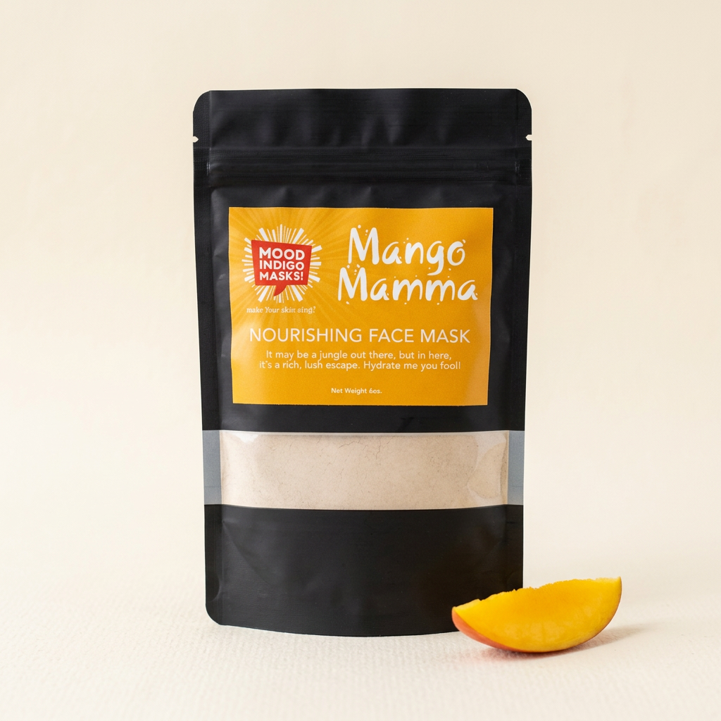 Mood Indigo Mango Mamma nourishing face mask in a black pouch with an orange label, beside a fresh mango slice on an ivory background.