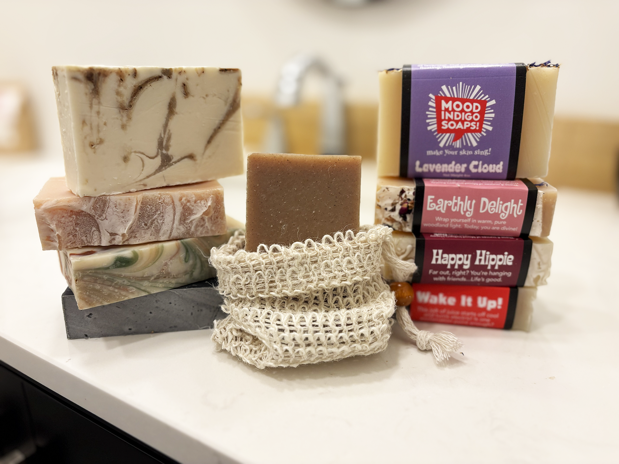 A group of handmade soaps and two reusable cotton soap suds bags arranged on a white countertop. The soaps include marbled cream, brown, and green bars, a solid medium brown bar, and packaged bars labeled Lavender Cloud, Earthy Delight, Happy Hippie, and Wake It Up!.