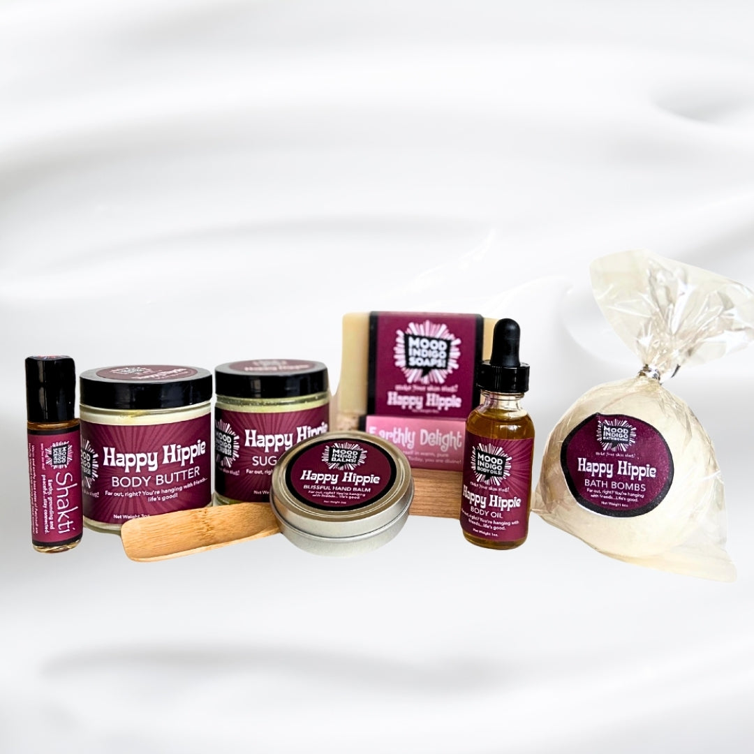 A curated wellness Patchouli gift set featuring Happy Hippie brand products, including body butter, body oil, a soap bar, a packaged bath bomb, a wooden spatula, and a small roll on perfume bottle, arranged on a white fabric background. Made by Mood indigo as part of their Earthy and Grounding collection, Happy Hippie