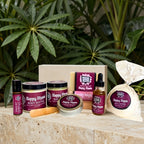 A curated wellness gift set from Natural Healing Apothecary Mood indigo. Arranged on a stone surface with green foliage in the background. The set includes a Happy Hippie body butter jar, a Happy Hippie hand balm tin, a Happy Hippie body scrub jar, a Happy Hippie essential oil roll on perfume bottle, a Happy Hippie soap bar, a Happy Hippie body oil, and a wood soap dish.