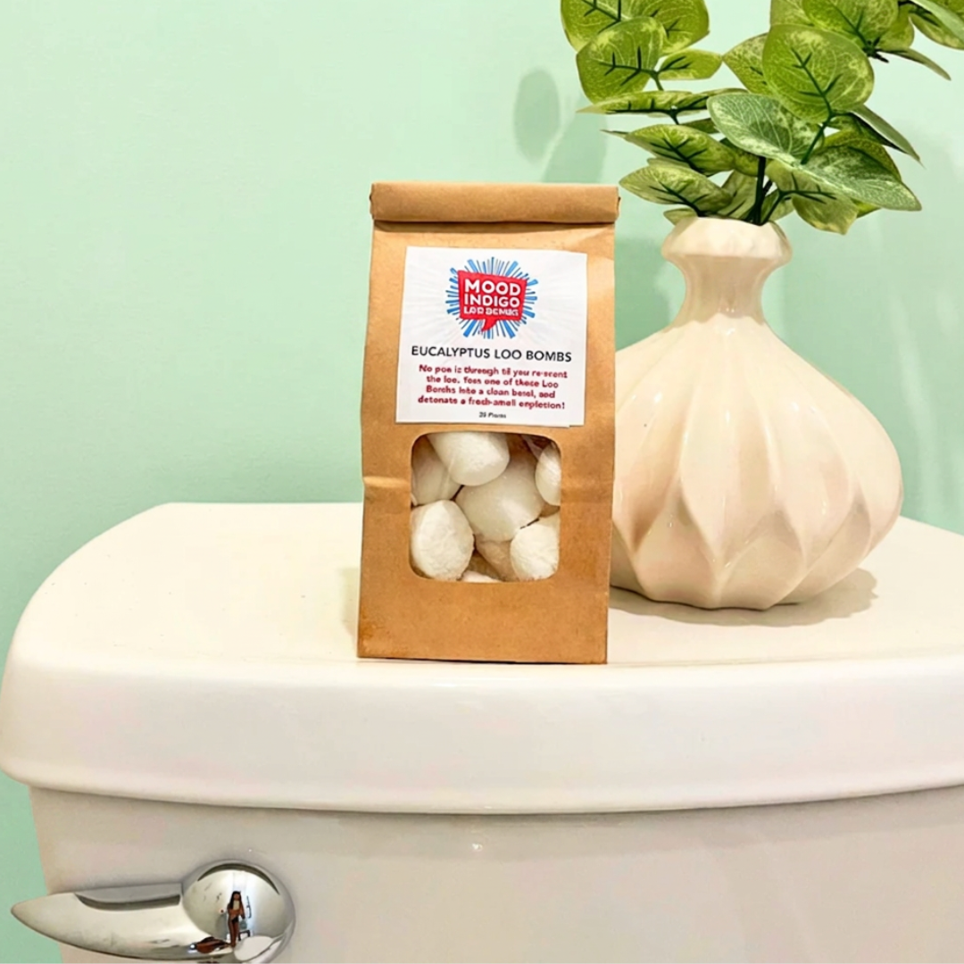 Mood Indigo Eucalyptus Loo Bombs packaging on a toilet with a plant in the background.E ucalyptus Potty Bombs for your potty. Clean and de-oderize your toilet with these essential oil toilet fizzies