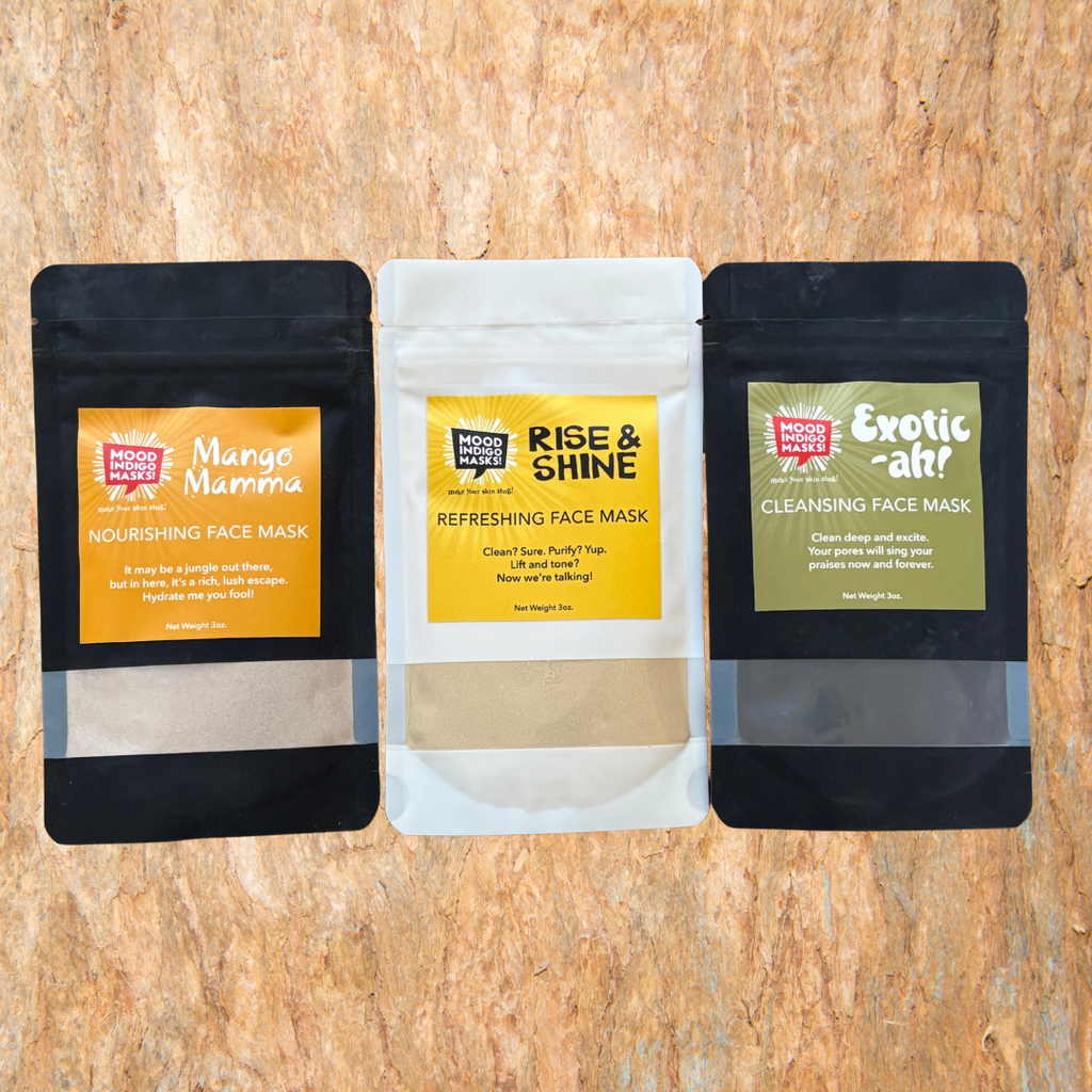 Three clay face mask packages by mood Indigo on a wooden surface. Mango Mamma, Rise & shine, and Exotic-ah!