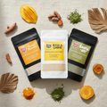Three stand-up pouches of Rise & Shine, Exotic-ah! and Mango Mamma  face masks arranged on a light fabric background with turmeric roots, dried leaves, and spice powders around them. The pouches are labeled Refreshing Face Mask, Cleansing Face Mask and Nourishing Face Mask. Made with a variety of dry, powdered clays and rice powder. 