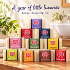 Ten Mood Indigo soap bars in colorful packaging stacked on a sunlit bathroom counter beneath “A year of little luxuries” and “Annual Soapscription.”