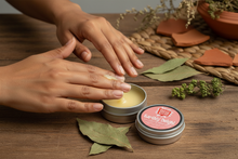 Woman applying EARTHLY DELIGHT Nourishing Hand Balm with shea butter, beeswax, and almond oil in natural earthy spa setting with patchouli botanicals and wooden textures
