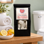Mood Indigo Citrus Loo Bombs 25-pack on an oak bathroom shelf beside lemons, orange peel and rolled white towels.