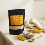Mango Mamma face mask pouch beside a ceramic bowl of mixed clay mask, a wooden spoon, cream towel, and mango slices on a sunlit vanity.